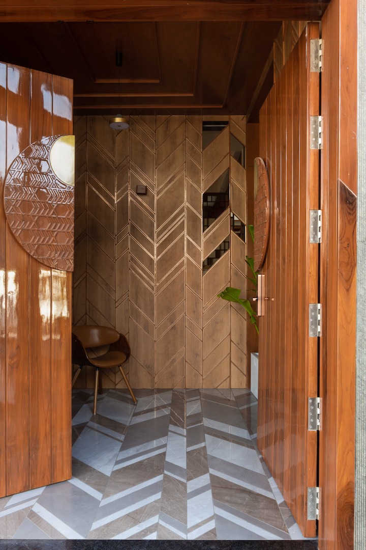 Teak pivot doors with double-height chevron-patterned feature wall — the same geometry continuing into the foyer floor tiles